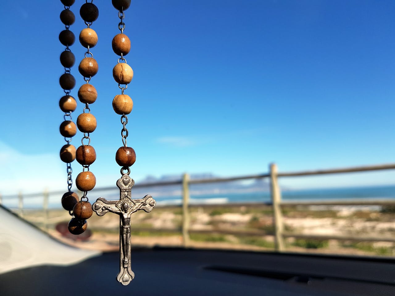 A wooden rosary with a crucifix hangs inside a car, overlooking a scenic ocean view.